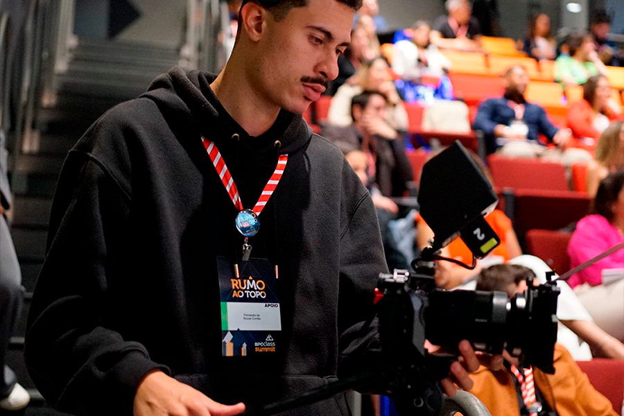 Homem centralizado, com câmera de vídeo, fazendo produção de vídeo, atrás dele, cadeiras de auditórios laranjas e pessoas sentadas
